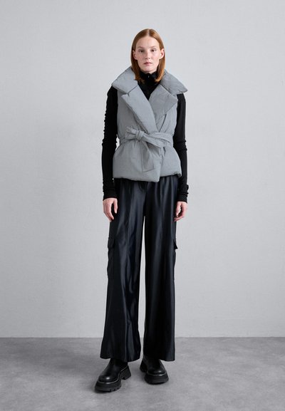 Ljusgrå vadderad väst med bred krage, belagd midja, bärs över en svart långärmad topp, tillsammans med svarta vida byxor och kraftiga stövlar.