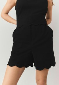 Top con orlo a scallop nero abbinato a pantaloni corti neri coordinati. Il tessuto appare liscio con tasche laterali e una vestibilità rilassata.