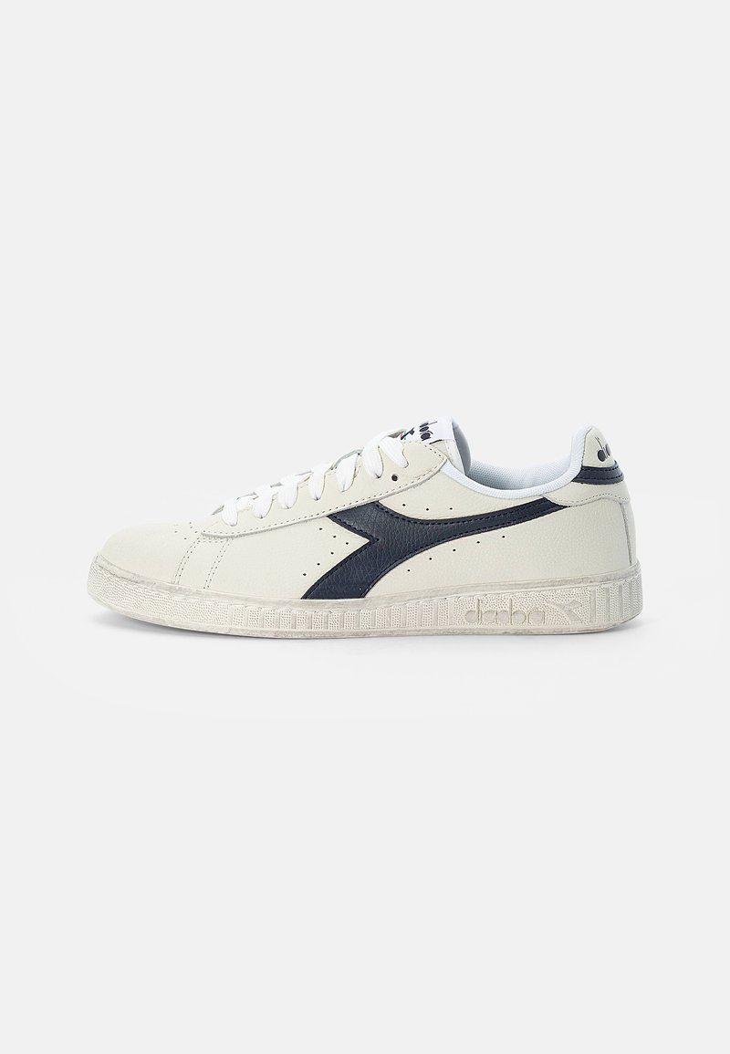 Zapatilla de piel blanca con detalles en azul marino, puntera redonda, suela de goma texturizada y lados perforados. Presenta cordones blancos y un logotipo.