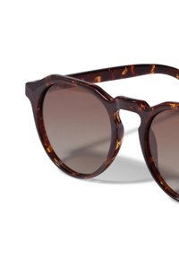 Gafas de sol de carey marrón con montura redondeada, lentes oscuros y una línea de cejas ligeramente puntiaguda. Textura suave y hardware mínimo.