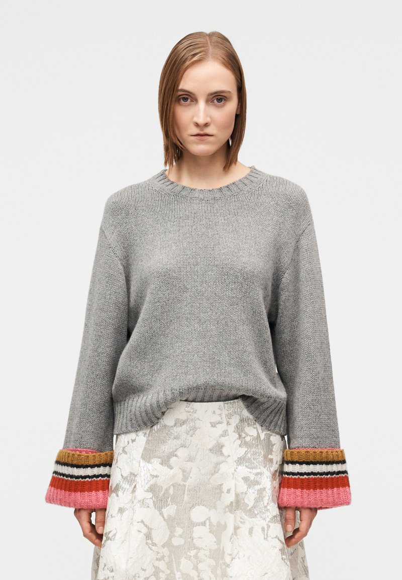 Pilkas megztas megztinis ilgomis rankovėmis su daugiakoliais dryžuotais rankogaliais. Derinamas su blizgia raštuota suknele.