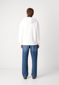 Felpa bianca con cappuccio e vestibilità comoda, abbinata a jeans larghi blu, su uno sfondo neutro, che mette in mostra un outfit informale.