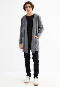Joven de pie, vistiendo un cárdigan con capucha gris, camiseta negra, pantalones negros y zapatillas blancas, contra un fondo blanco liso.