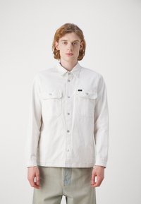 Camisa de mezclilla blanca de manga larga, con dos bolsillos en el pecho, cierre frontal abotonado y etiqueta de marca sutil. Cuenta con un cuello clásico y botones plateados.