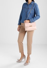 Světlounce růžová kožená kabelka s klopou a zlatými kovovými doplňky, nošená přes rameno. Kombinována s džínovou košilí a béžovými kratšími kalhotami.