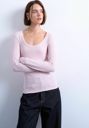 Femme aux cheveux bruns courts, portant un haut à manches longues rose clair et un pantalon ample foncé, debout les bras croisés devant un fond uni.