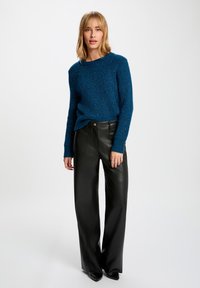 Blauwe gebreide trui met een textuurpatroon, gecombineerd met hoge zwarte leren wijde broek. De outfit heeft een casual, relaxte pasvorm.