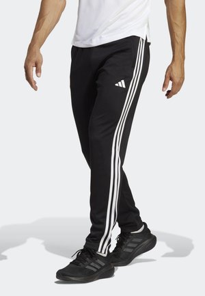 Mężczyzna ubrany w czarne spodnie dresowe Adidas z białymi paskami i czarne trampki, stojący z lekko skrzyżowanymi nogami.