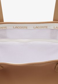Tan Lacoste tote táska belseje, fehér hálós rekesszel és sima barna béléssel. Arany logó nyomtatva a textil díszítményen.