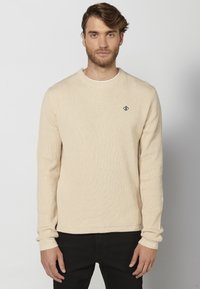 Beige geribbelde trui met lange mouwen, ronde halslijn en subtiele logo borduursel. Draagt over zwarte broek, met een gestructureerde stofafwerking.