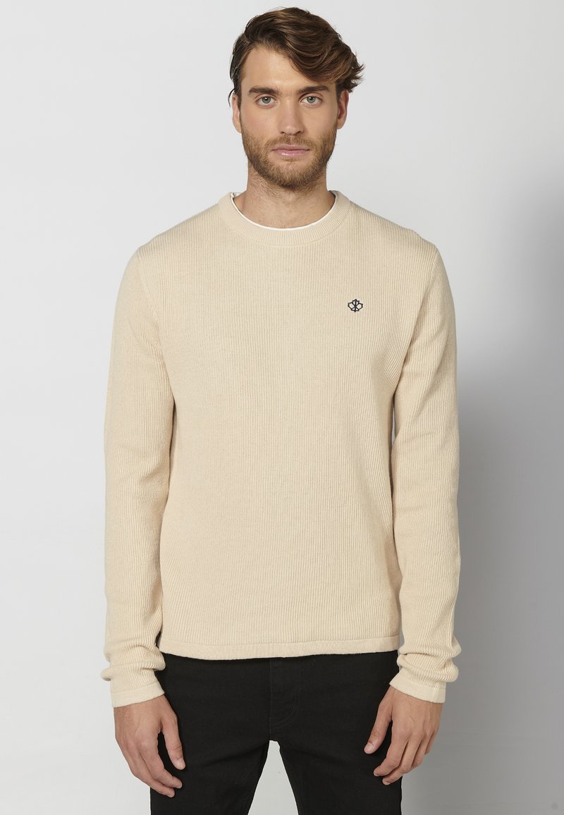Beige geribbelde trui met lange mouwen, ronde halslijn en subtiele logo borduursel. Draagt over zwarte broek, met een gestructureerde stofafwerking.