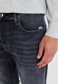 Jeans din denim gri, cu croi slim, având un buzunar frontal, textură subtilă și etichetă G-Star pe buzunar. Mână arătată odihnindu-se pe jeans.