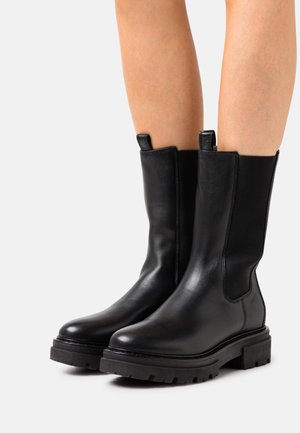 Bottes en cuir noires mi-mollet avec des semelles épaisses et des panneaux latéraux élastiques portées sur des jambes nues sur un fond clair.