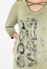 Olivgrünes Oberteil mit langen Ärmeln, U-Ausschnitt und Kreuzungsdetails. Mit metallic silbernem Druck und Pailletten in verschiedenen Mustern.