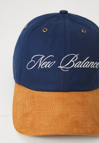 Navy blue cap with a brown suede visor, featuring embroidered "New Balance" text. Metal ventilation grommets on the crown.