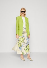 Limoengroene textured blazer over een witte top, gecombineerd met een crème rok met bloemenpatroon en lichtbeige puntige hakken. Houdt een beige tas vast.