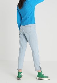 Personne portant un jean bleu clair avec des détails lacés aux chevilles, des baskets montantes vertes et un pull texturé bleu vif, debout de dos.