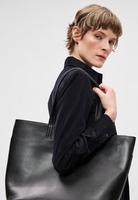 Černá kožená taška přes rameno s hladkou texturou, širokým otevřeným tvarem a dlouhými uchy, držená osobou v tmavé košili na knoflíky.