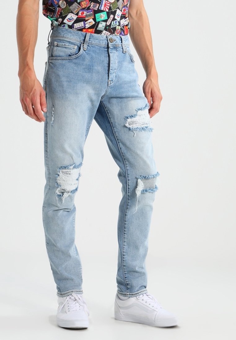 Jean en denim bleu clair avec des déchirures et des bords effilochés, présentant une coupe slim et un design cinq poches, associé à des baskets blanches.