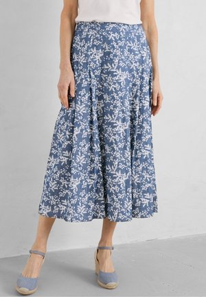 Femme portant une jupe midi fleurie bleue avec des motifs blancs et des chaussures compensées bleues à bride à la cheville, debout sur un fond uni gris clair.