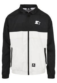 Giacca a vento in bianco e nero con zip frontale, colletto rialzato, polsini elasticizzati e tasche laterali. Presenta il logo sul petto e sul pannello superiore.