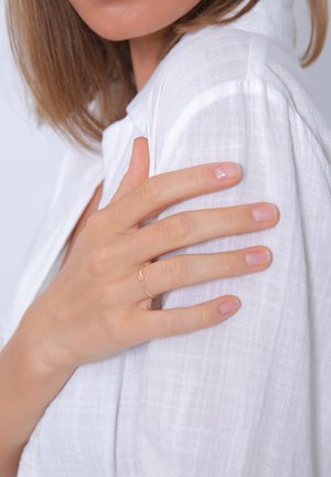 Goldring an einer Hand, vor einem weißen, strukturierten Hemd. Der Ring hat einen kleinen Akzentstein. Die Hand hat natürliche, unpolierte Nägel.