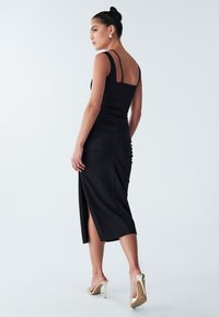 Black midi dress with a fitted silhouette, ruched detailing, thin straps, and a side slit, paired with gold high-heeled sandals.