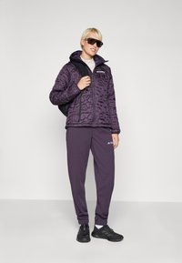 Lila quiltad jacka med huva, dragkedjestängning och logotyp. Kombinerad med matchande lila joggers och svarta sneakers, accessoriserad med solglasögon.