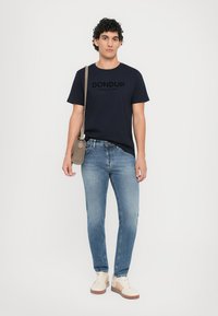 Navy cotton t-shirt featuring "DON DUP" logo, paired with light blue jeans. Model wears cream sneakers and carries a small brown bag.