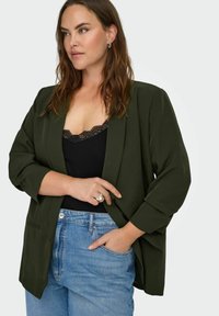 Sieviete ar gariem brūniem matiem, tērpusies tumši zaļā žaketē pār melnu topu ar mežģīņu apmali un zilās džinsās, pozē ar vienu roku kabatā, redzami gredzeni.