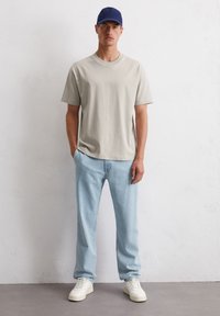 Lichtgrijze katoenen t-shirt, ruime pasvorm, gecombineerd met lichtblauwe denim jeans en witte sneakers. Ook een marineblauwe pet dragend, staand op een grijze ondergrond.