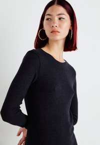 Ribbed long-sleeve top in dark gray with a round neckline. Smooth texture and fitted design highlight the silhouette. Large hoop earrings visible.