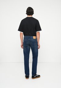 Camiseta negra con ribete blanco, jeans de mezclilla azul y zapatillas negras con suelas marrones. Parche de logo visible en la cinturilla trasera de los jeans.