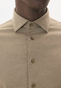Šviesiai rudas raštuotas marškinis su klasikiniu apykakle, penkiais mygtukais ir tekstūruotu audiniu su subtiliais tinklo raštais.