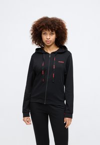 Svart zip-up hoodie av mjukt tyg, med huva, två framfickor och röda logotypdetaljer på bröstet och dragskor.