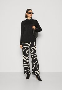 Zwarte button-up shirt met kraag, gecombineerd met hoge taille, wijdvallende broek met een abstract wit patroon. Zwarte puntige laarzen en tas.