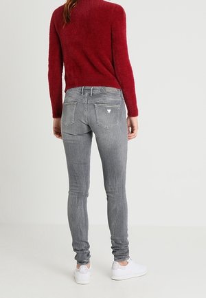 Vrouw staand, gekleed in nauwsluitende grijze spijkerbroek met achterzakken en een rode trui met lange mouwen, gecombineerd met witte sneakers, tegen een effen achtergrond.