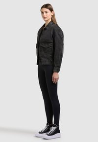 Fekete farmerdzsekit, fekete leggingset és fehér fűzős, fehér talpú fekete magas szárú tornacipőt viselő, oldalról látható nő.