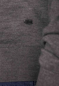 Grijs geribbelde trui met een zwart logo label, met een close-up van de stofstructuur en de afwerking van de manchetten.