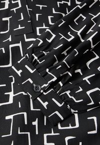 Camicia di tessuto nero con motivi astratti bianchi, dotata di polsini con bottoni e dettaglio arricciato sulle maniche. Texture liscia.