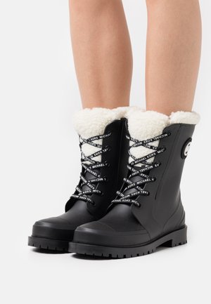 Svarta vattentäta stövlar med vit fuskpälsfoder, robust sula och detaljerade snören med varumärkesnamn. Slät yta, midja till vaden.