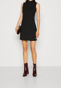 Robe noire sans manches à col montant, agrémentée d'accents de ferrures dorées à la taille. Associée à des bottines bordeaux brillantes.