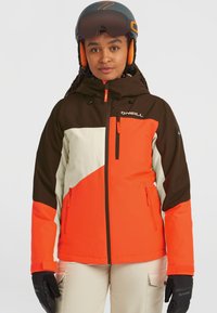 Veste de ski colorée avec des panneaux orange, marron et crème. Fabriquée en matériau synthétique avec une fermeture éclair à l'avant et une capuche. Gants noirs.