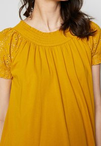 Blusa gialla senape con un collo arricciato testurizzato e maniche corte a sbuffo con dettagli in pizzo. Tessuto morbido e leggero.