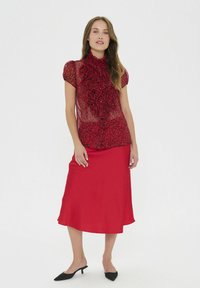 Blusa roja transparente con cuello con volantes sobre una falda de satén roja lisa. El conjunto cuenta con mangas cortas y zapatos planos negros con punta.