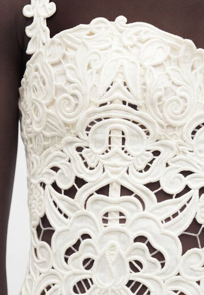 Λευκό δαντελένιο ύφασμα με περίπλοκα λουλουδένια και σπειροειδή σχέδια πάνω σε σκούρο δέρμα, που αναδεικνύει λεπτομερή κέντημα και αποκομμένα μοτίβα.