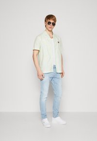 Camisa verde claro de manga corta con un patrón sutil, camiseta blanca, jeans azul claro y zapatillas blancas. Accesorios: gafas de sol.