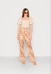 Mulher vestindo uma blusa bege de mangas bufantes, calças largas com padrão em espiral laranja e bege, sapatos de salto aberto laranja e óculos de sol laranja, de pé contra uma parede branca.