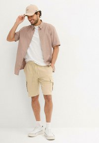 Kurzärmliges, hellrosa Hemd mit Knöpfen über einem weißen T-Shirt, beigen Cargo-Shorts und weißen Sneakers mit beigen Akzenten, dazu eine passende Kappe.