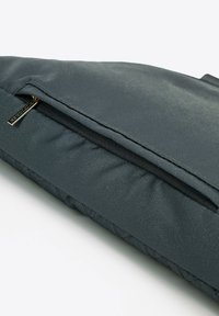 Šedá polstrovaná taška s texturovaným, hladkým povrchem, obsahující kapsu na zip a malý zlatý detail emblému na boku.